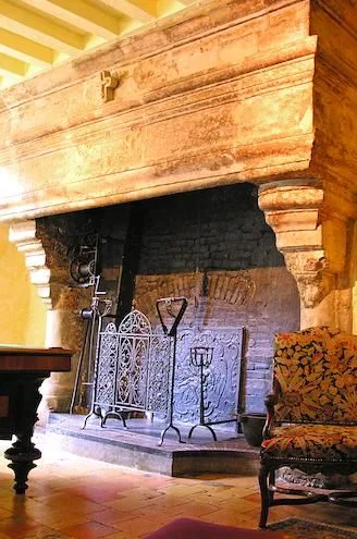 Monumental stone fireplace with carved corbels and wrought-iron fire screens, a tapestry armchair beside it