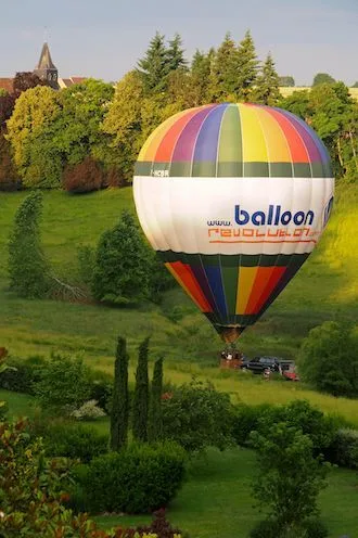 Rainbow-striped hot-air balloon coming down in a green valley, a church spire beyond the trees