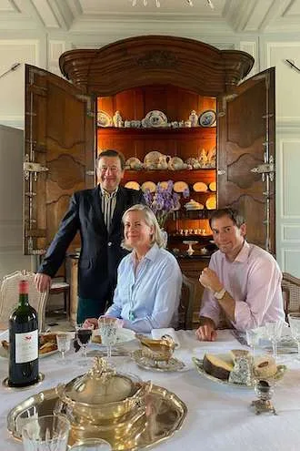 Two men and a woman at a laid table before an open oak dresser filled with porcelain