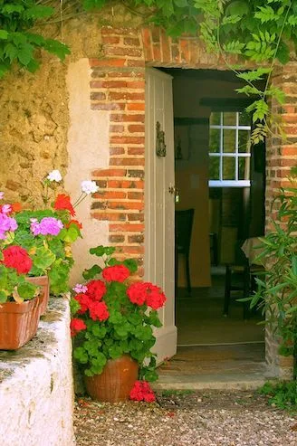 Open cottage door framed in old brick, with pots of red geraniums on the step