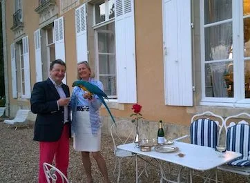 A man and a woman holding a green parrot on the terrace, champagne and a rose on the garden table