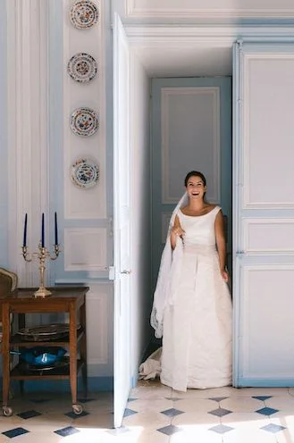 Smiling bride with a long veil stepping through a doorway, a candelabra and wall plates beside her